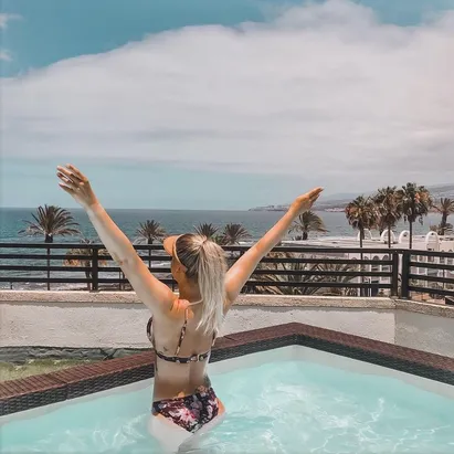 Blonde woman in a swimsuit with her hands raised, looking out towards the ocean from the H10 Conquistador swimming pool.