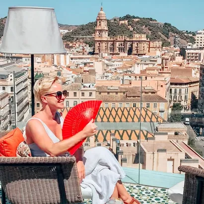 Donna con capelli corti seduta sulla terrazza dell'H10 Croma Málaga, con in mano un ventaglio e la vista sulla città sullo sfondo.