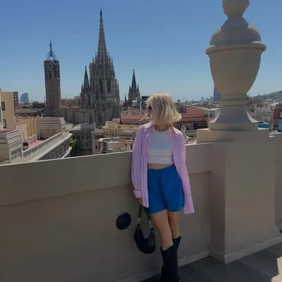 Chica rubia de pelo corto con camisa ovsersized rosa, tejanos cortod y botas negras en el rooftop de H10 Madison con las vistas de la catedral de Barcelona